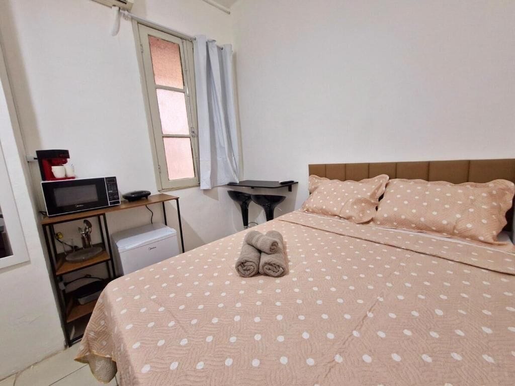 A bedroom with a bed, pillows, a folded towel, a desk with a computer, a microwave, and a small refrigerator.