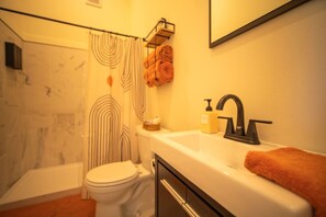 Une salle de bain avec des toilettes, un lavabo doté d’un robinet noir, un flacon de savon et une serviette accrochée au mur.