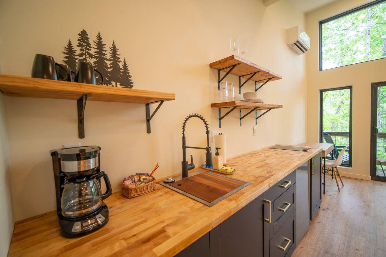 A modern kitchen with wooden countertops, a coffee machine, and a sink. There are shelves above the counter holding glasses and a decorative tree.