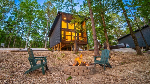 Una cabaña con un hogar a la vista y dos sillas Adirondack en un área boscosa.