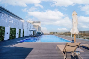 A rooftop pool area with a clear blue pool, lounge chairs, and a white building with a geometric facade.