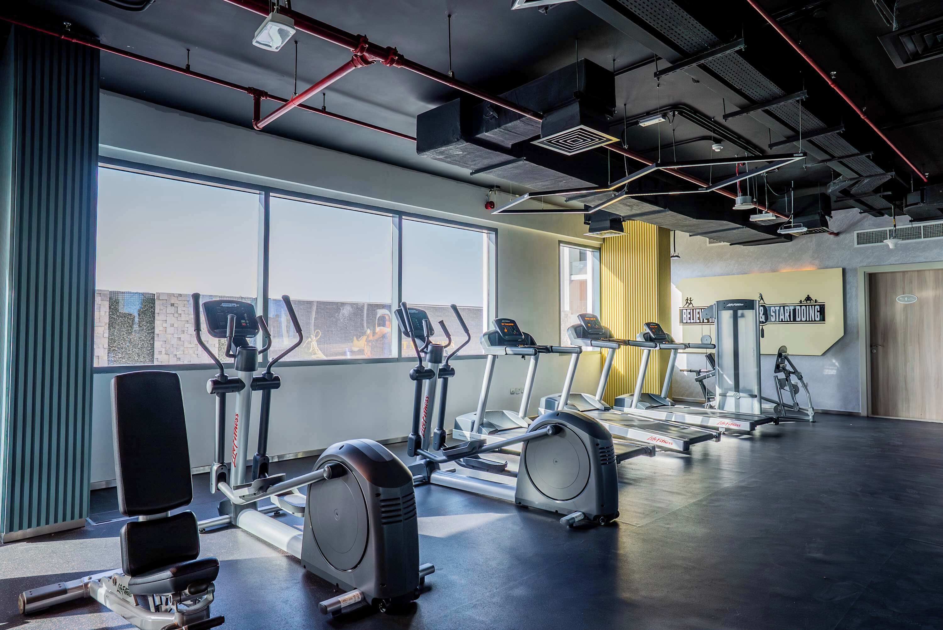 Une salle de sport équipée de divers appareils d’exercice, dotée de grandes fenêtres et d’un plafond au design industriel moderne.