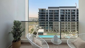 A balcony with a view of a modern building, a swimming pool, and two white wicker chairs.