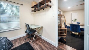 A compact office space with a glass desk, a chair, a wicker chair, and a dining area with a table and chairs.