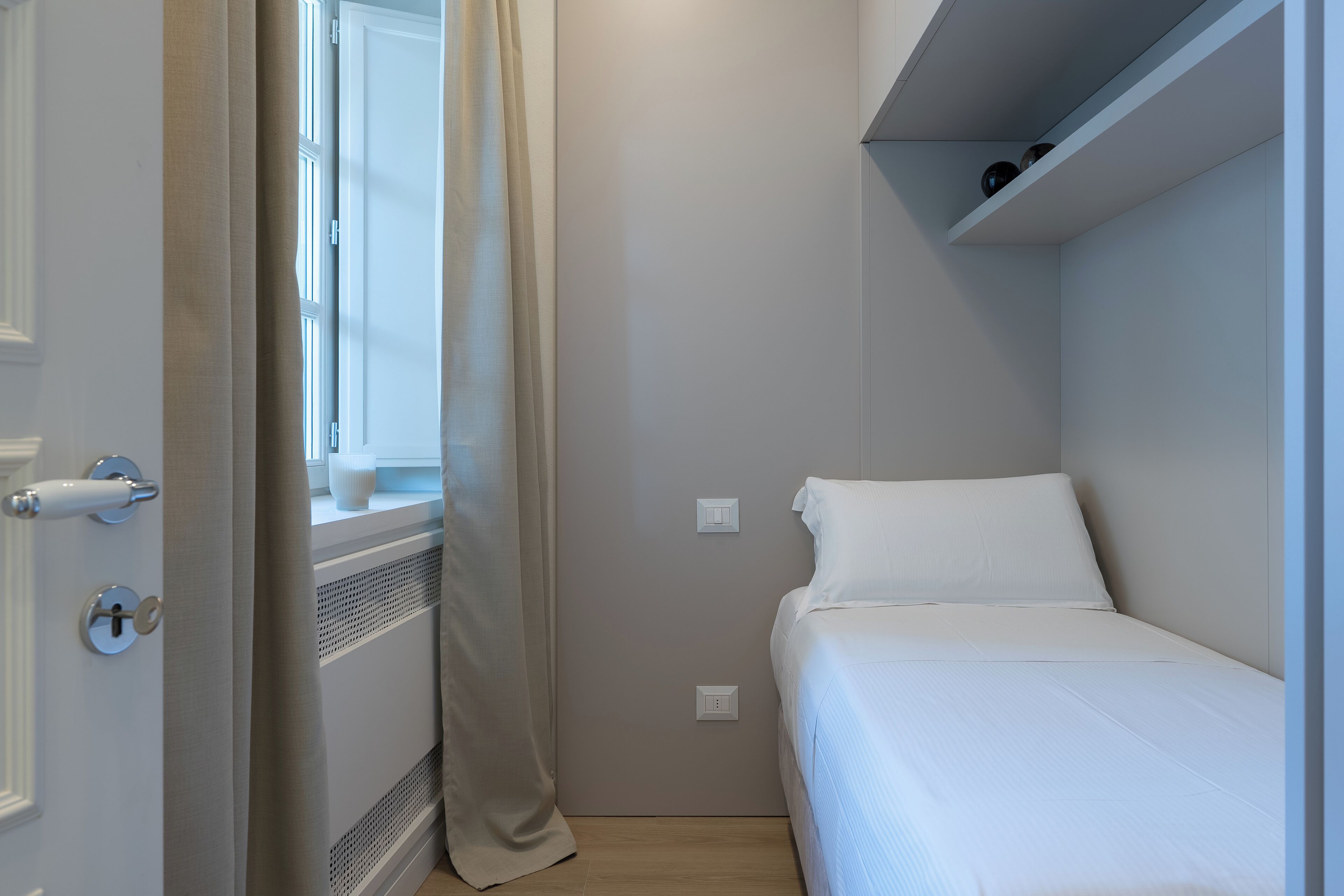 Un dormitorio pequeño y ordenado con una cama individual, una ventana con cortinas, un radiador y un estante sobre la cama.
