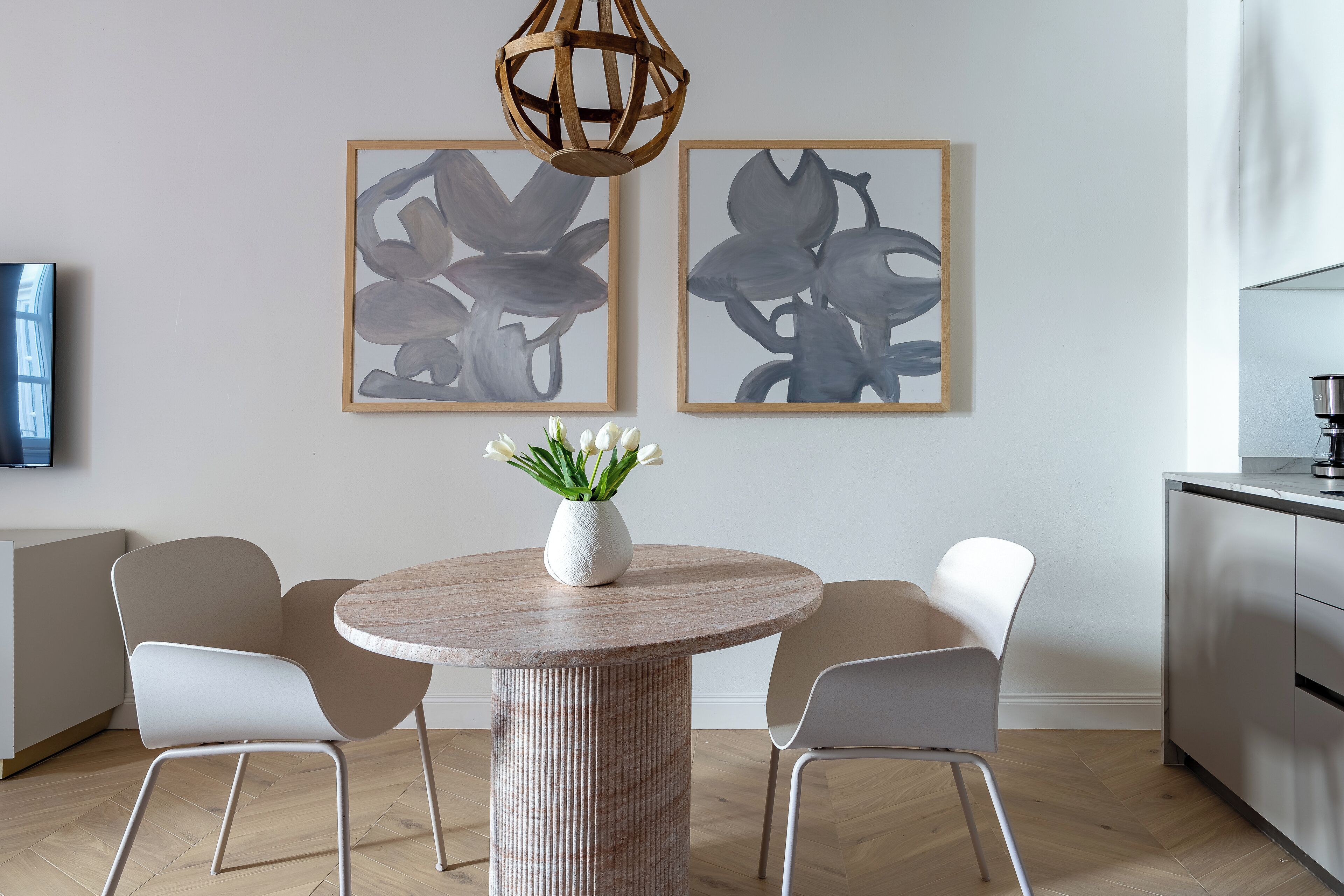 A modern dining area with a round table, two chairs, a vase with flowers, and two framed artworks on the wall.