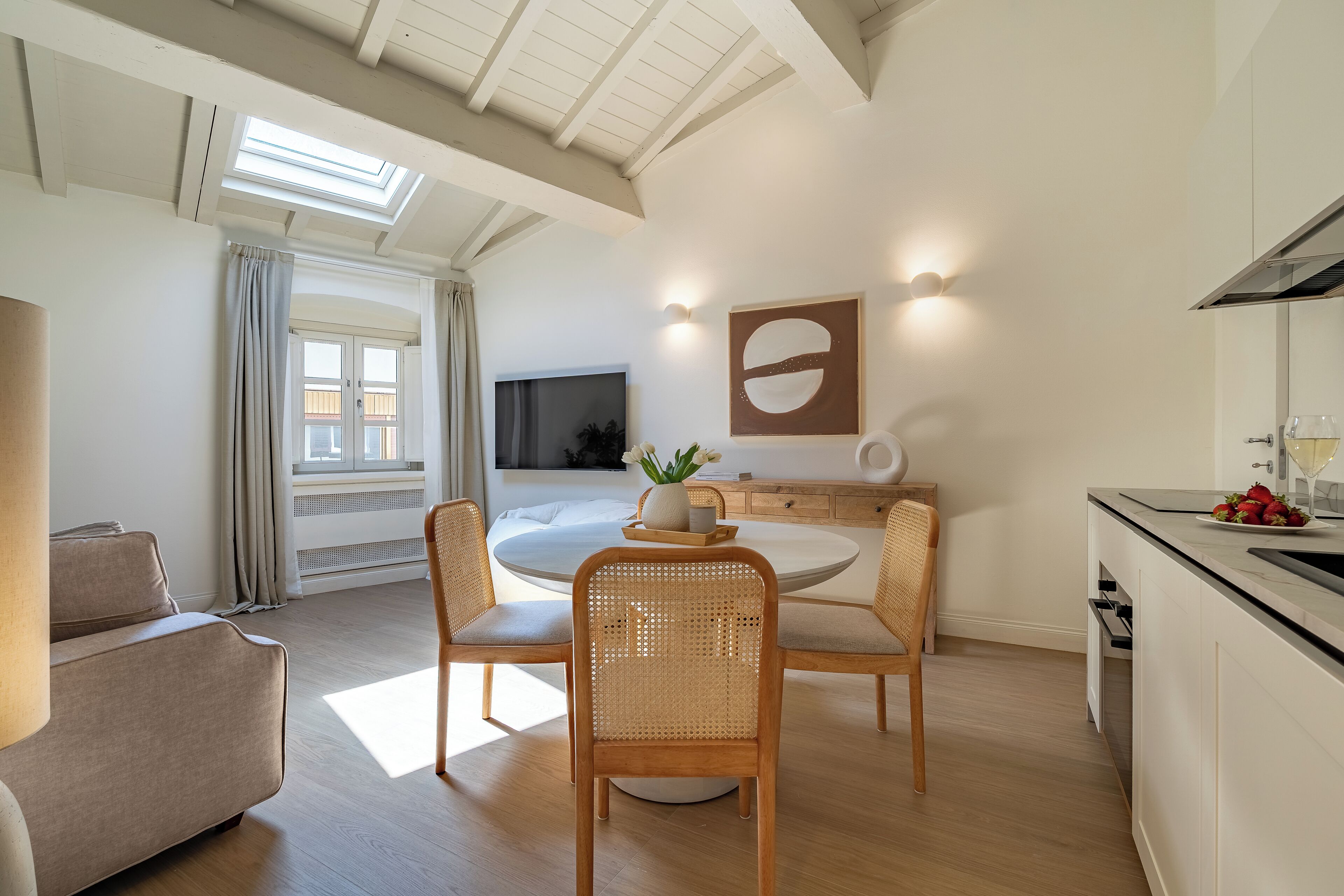 A modern living room with a round dining table, wicker chairs, a sofa, and a kitchen area with a dining table and chairs.