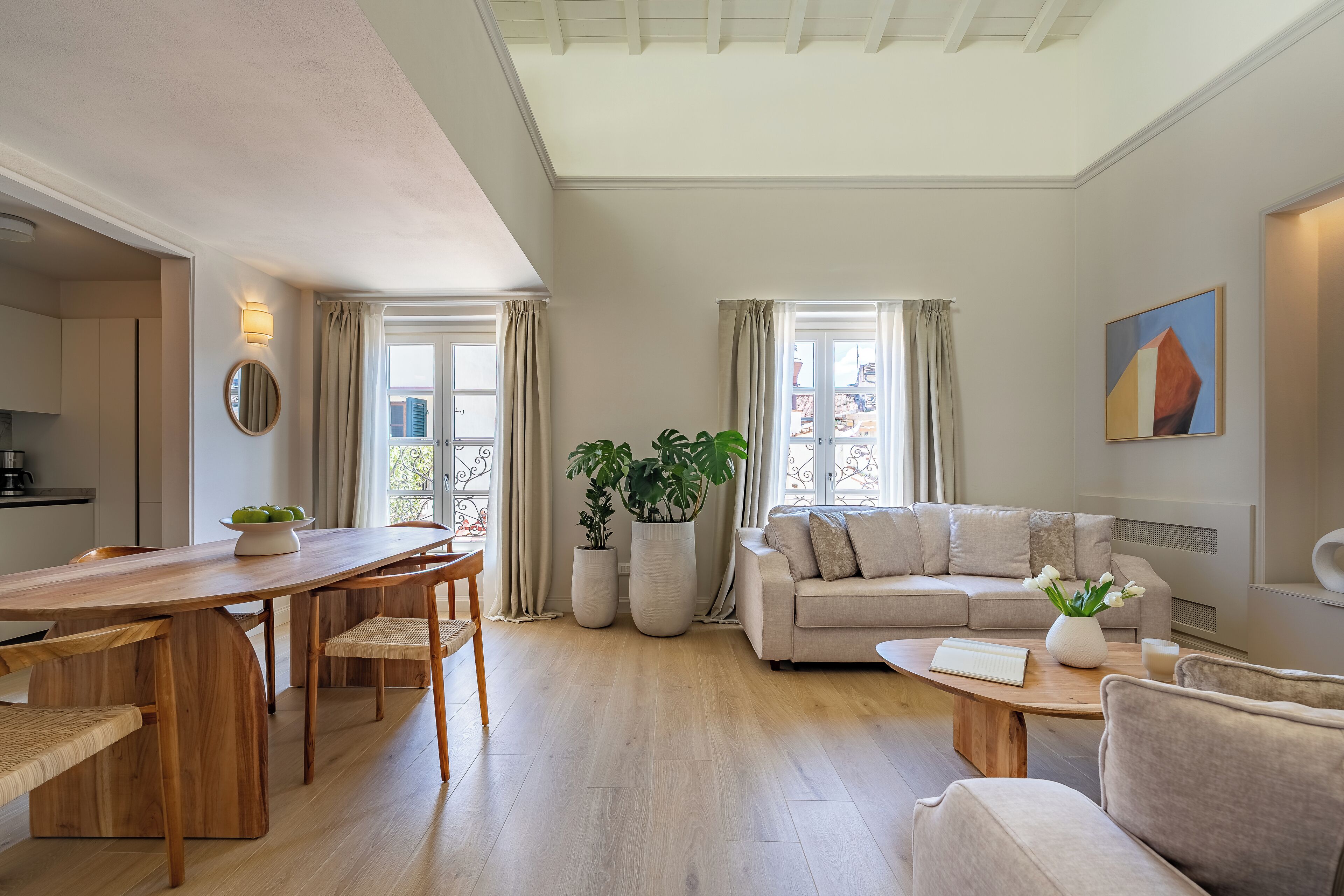 A modern living room with a wooden dining table, beige sofa, and large windows with curtains.