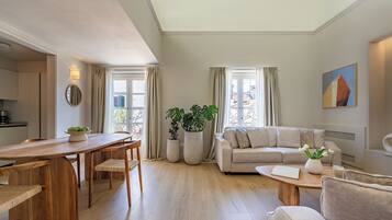 A modern living room with a wooden dining table, beige sofa, and large windows with curtains.