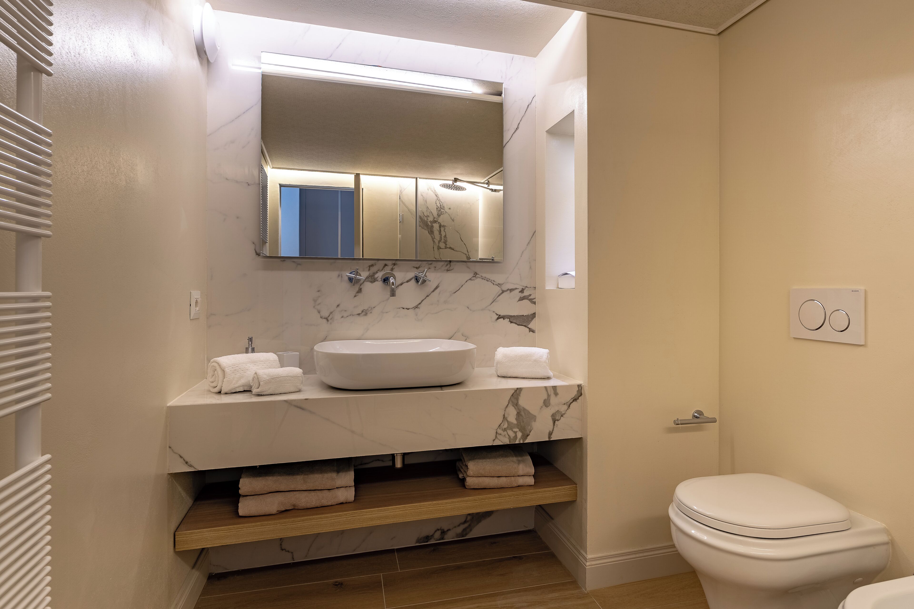 Un baño moderno con lavamanos de mármol, un espejo encima y un inodoro. Hay toallas sobre el lavamanos y un estante debajo.