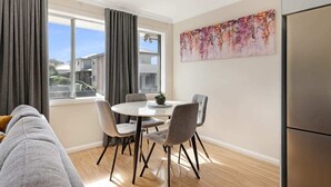 Una sala moderna con un sofá gris, una mesa de comedor con cuatro sillas, un ventanal con cortinas y un cuadro abstracto colorido en la pared.