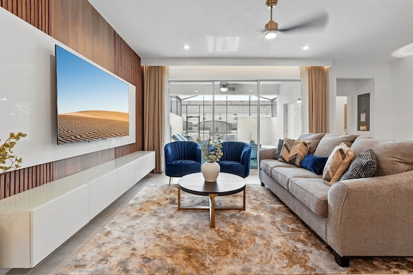 A modern living room with a large flat-screen TV, a sofa, a coffee table, and a view of the outside through large windows.