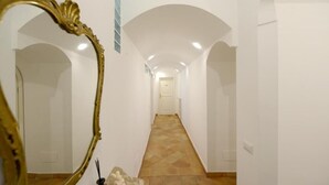 Un couloir aux murs blancs et au sol carrelé, qui mène à une porte au fond.