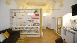 Un espace de vie compact comprenant un canapé, une étagère blanche agrémentée d’objets décoratifs, une petite table ronde et un coin cuisine avec un miroir et des appareils électroménagers.