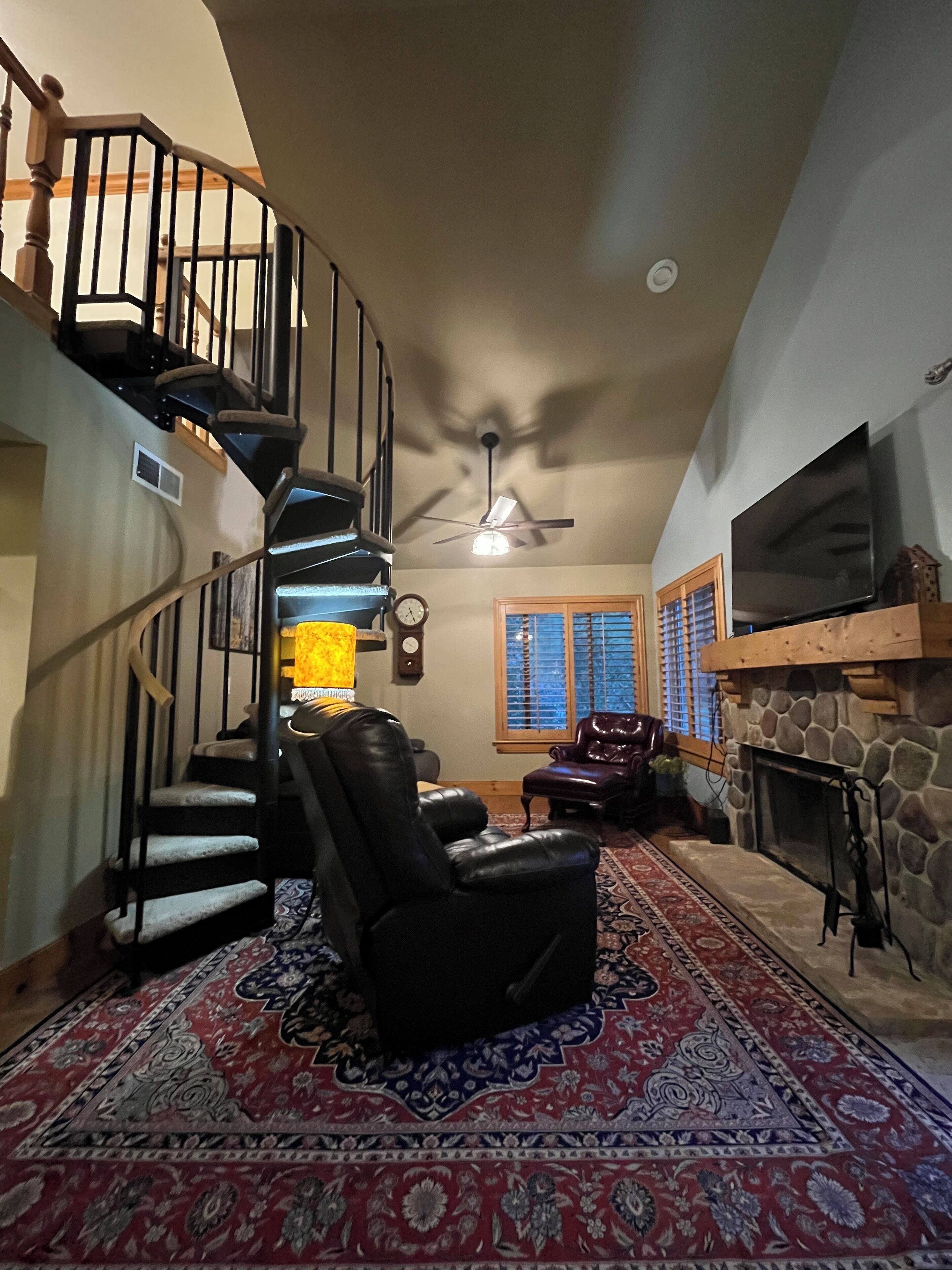 Un salon spacieux avec une cheminée, un grand téléviseur à écran plat, un fauteuil inclinable en cuir, un tapis à motifs et un escalier en colimaçon.