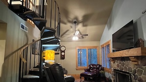 Amplia sala de estar con chimenea, televisor de pantalla plana grande, sillón de cuero reclinable, alfombra estampada y escalera espiral.