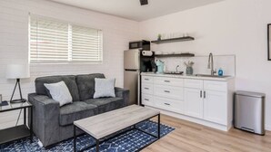 Un espace de vie compact comprenant un canapé gris, une table basse en bois, une kitchenette avec des meubles blancs et un ventilateur de plafond.