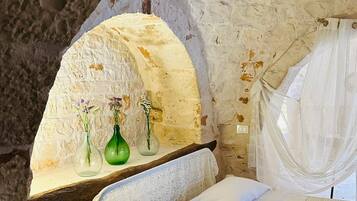 Una habitación rústica con pared de piedra, un banco de madera, una cama con colcha blanca y tres jarrones con flores.