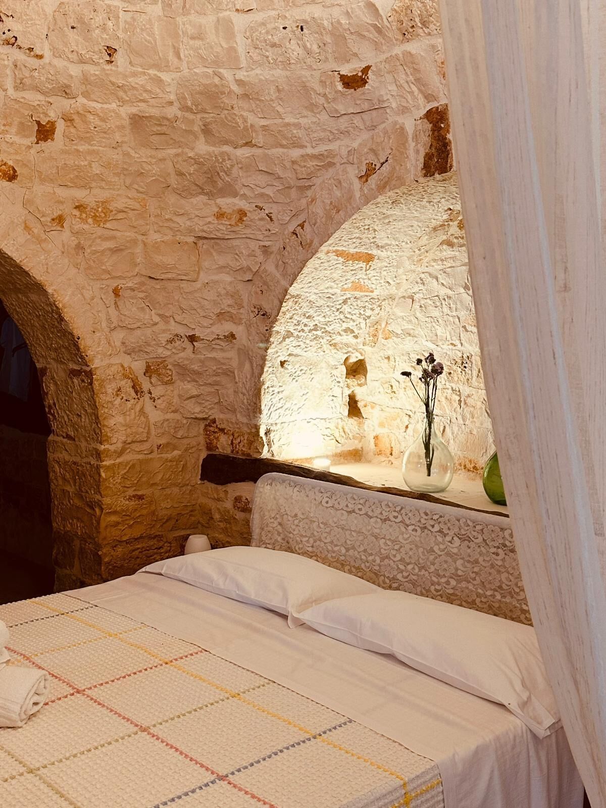 A room with a stone wall, a bed with a checkered bedspread, and a vase with dried flowers on a windowsill.