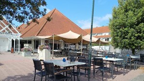 Ett utomhuskafé med bord och stolar, skyddat av parasoller, och en byggnad med ett tegeltak i bakgrunden.