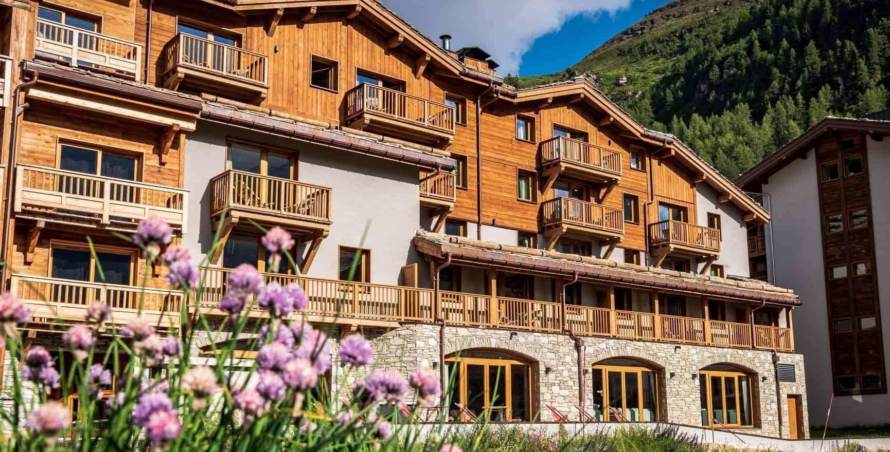 A multi-story wooden building with balconies, stone accents, and a mountainous backdrop.
