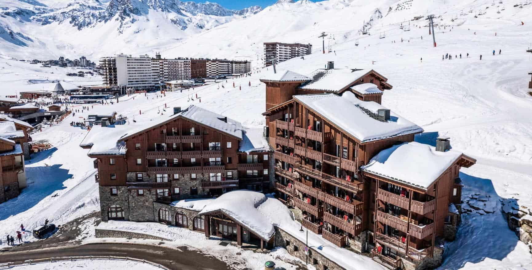 Une station de montagne enneigée, avec des bâtiments en bois, des pistes de ski et un ciel bleu dégagé.