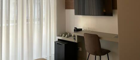 A modern hotel room with a bed, a desk, a chair, a television mounted on the wall, and a large window with sheer curtains.