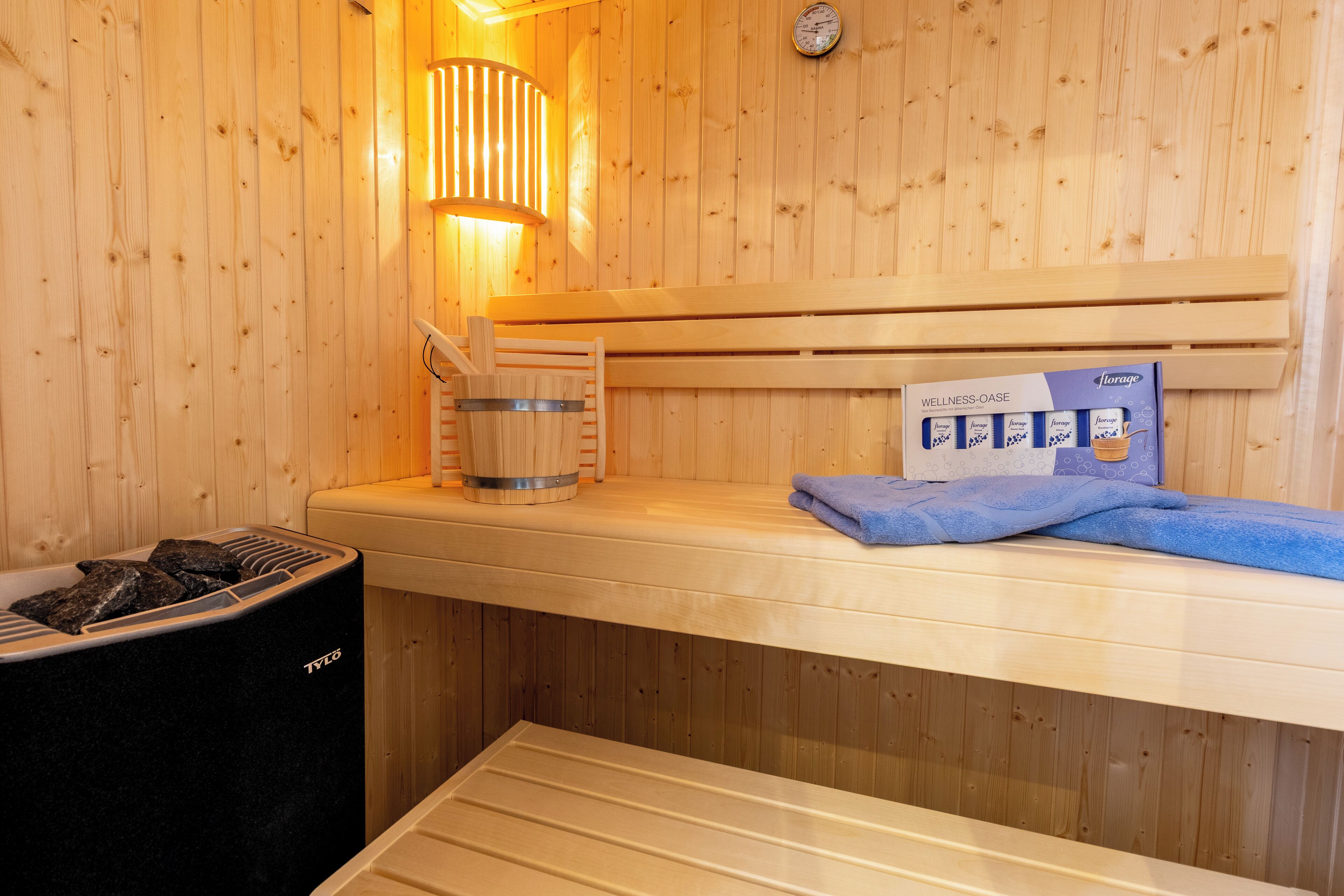 A sauna room with wooden walls, a bench, a towel, a bucket, and a storage box labeled "WELLNESS-OASE".