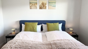 Una cama bien hecha con sábanas blancas y un suave plaid blanco, flanqueada por dos mesitas de noche con lámparas, una de ellas con un libro encima.