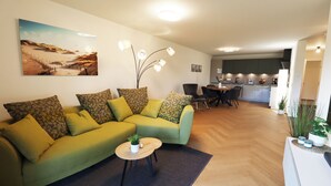Una sala de estar moderna con un sofá verde, una mesita redonda con una planta y un comedor al fondo.