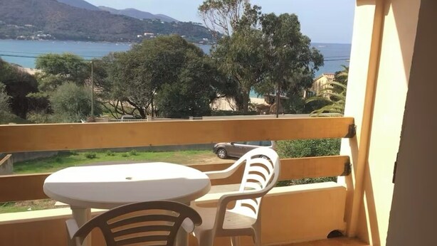 A balcony with a round table and two chairs, overlooking a scenic view of a coastal area with trees and hills.