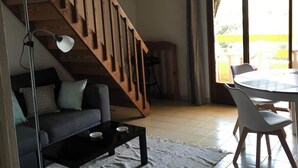 A living room with a sofa, coffee table, and a dining area with chairs and a table. A wooden staircase leads to an upper level.