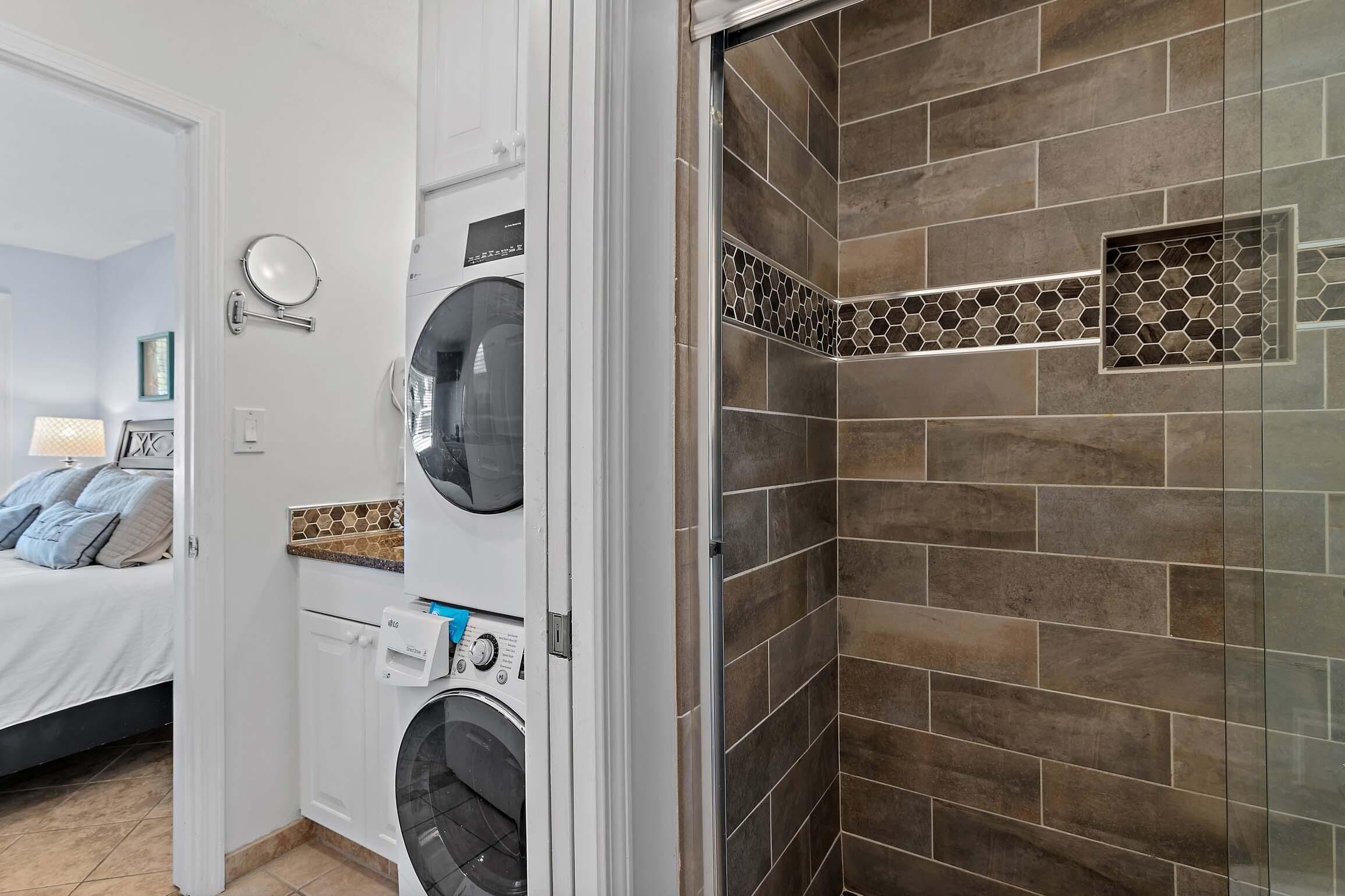 Uma área de lavandaria com máquina de lavar e secadora, um espelho e um chuveiro revestido com azulejos marrons.
