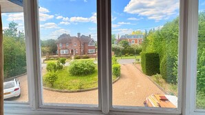 A view through a window of a residential area with a brick house, a driveway, and a well-maintained garden. - 6 Bed House on Prime Private Road in Ickenham (Ickenham)