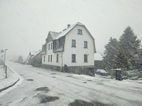 A snow-covered residential street with a white house, a parked car, and a snow-covered tree.
