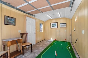 A room with a green artificial turf putting green, a wooden desk, a chair, and a door.