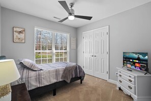 A bedroom with a bed, a TV, a ceiling fan, a window with blinds, and a door.