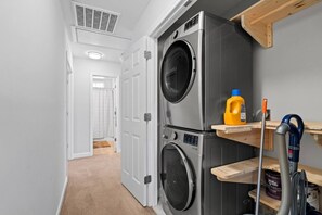 A laundry room with two stacked front-loading washing machines, a wooden shelf with cleaning supplies, and a vacuum cleaner.