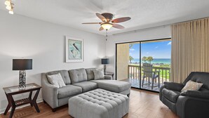 A modern living room with a sectional sofa, ottoman, and a black leather chair. A ceiling fan and two floor lamps are visible. The room has a large sliding glass door leading to a balcony with a view of the ocean. - Blue Surf 16B (Miramar Beach)