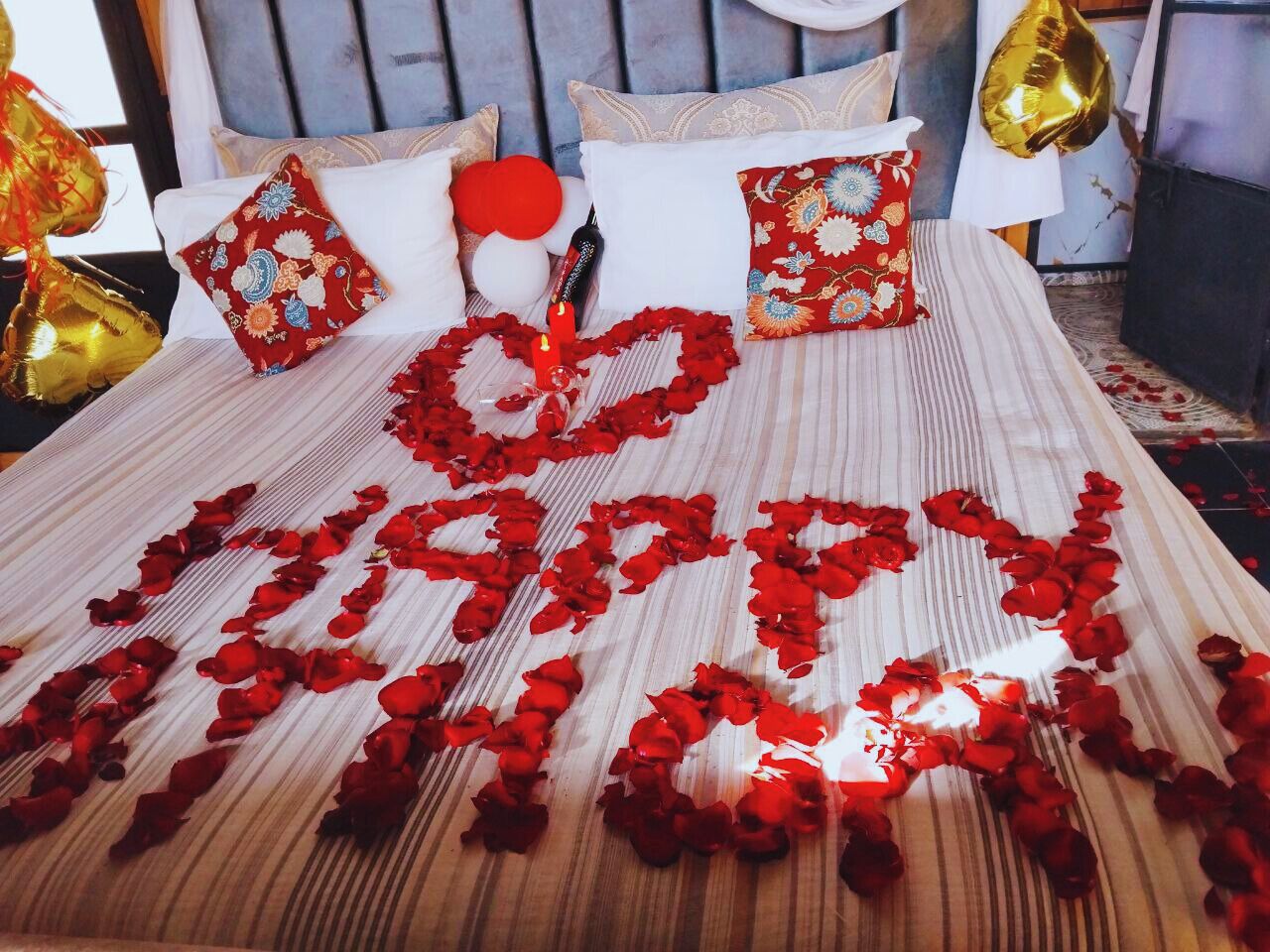 A bed with rose petals arranged to spell "LOVE" and a heart shape, surrounded by pillows and balloons.