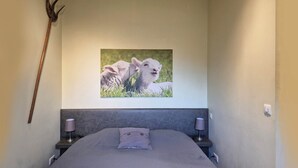 A bedroom with a bed, two bedside lamps, a wall-mounted picture of lambs, and a wooden staff leaning against the wall.