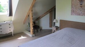 A bedroom with a sloped ceiling, a bed, a dresser, a chair, a lamp, and a painting on the wall. - Quiet Chambre des Daims (BERTHEN)