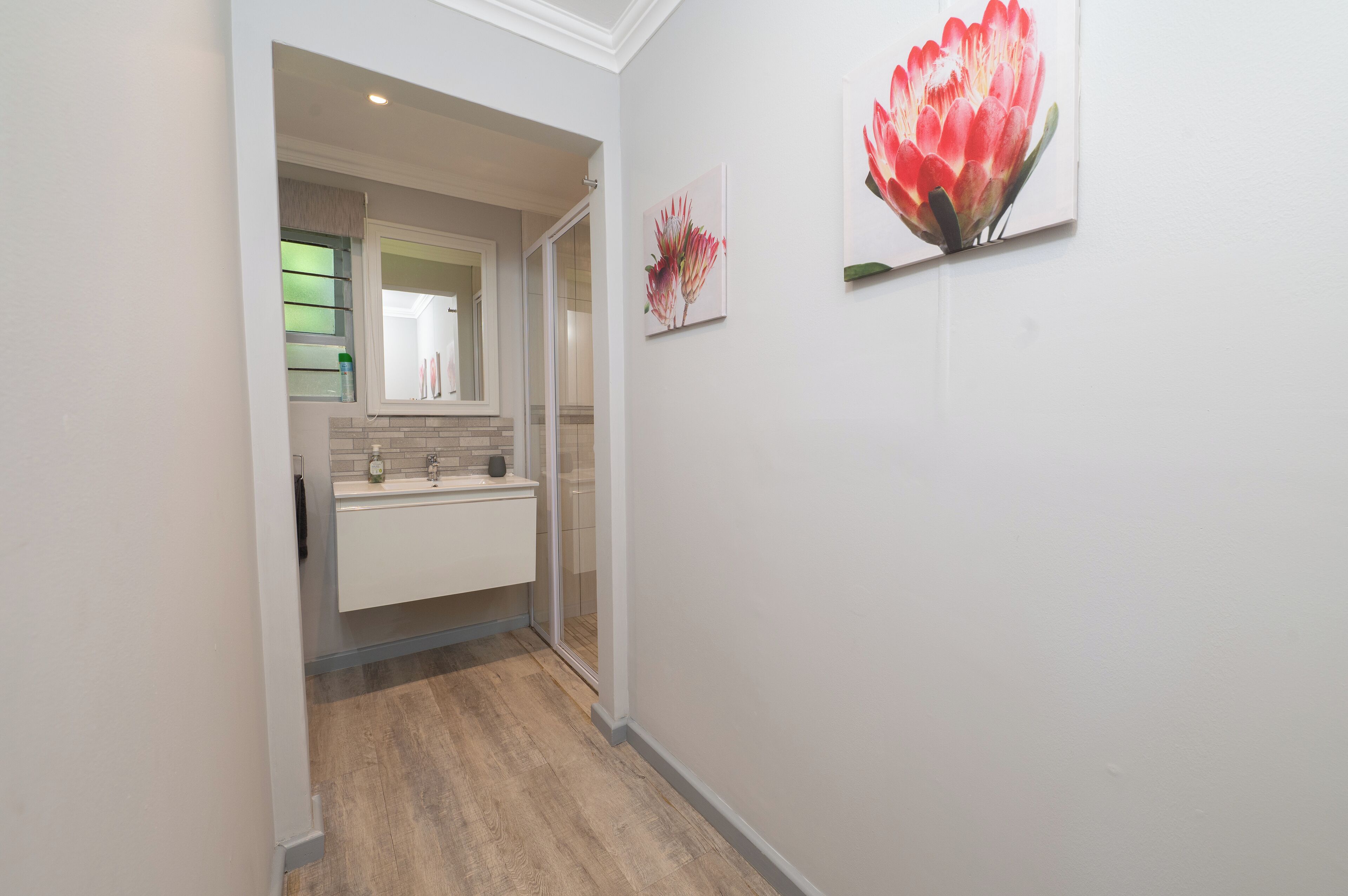 A hallway leading to a bathroom with a white sink, a mirror, and a door. There are two framed floral artworks on the wall.