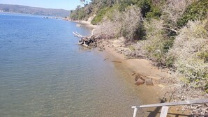A clear view of a beach with a wooden staircase leading down to the water, surrounded by dry vegetation and a distant view of hills.