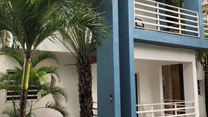 A modern two-story building with a balcony and a small garden area with palm trees. - Cozy 1-bedroom apartment with AC, WiFi in beautiful Baní (Baní)