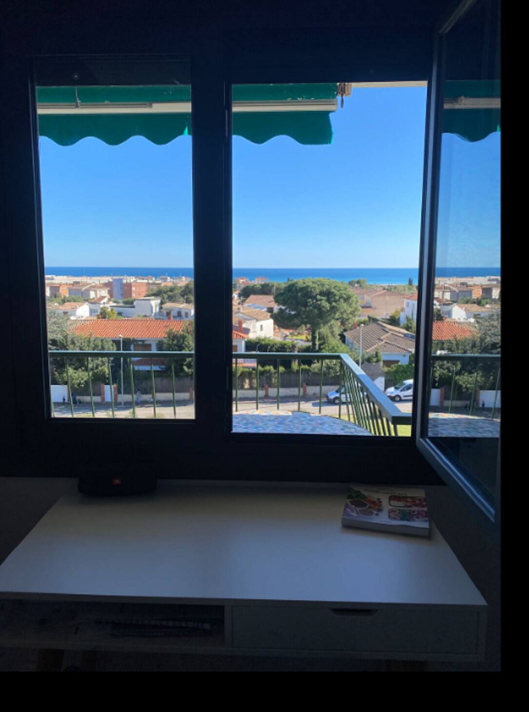 A room with a window overlooking a coastal town, a small table, and a book on the windowsill.
