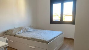 A single-bed room with a white bed, a small desk, and a window with blinds. - Bright apartment with sea view (Calafell)