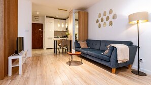 A modern living room with a sofa, a TV, and a lamp. There is a kitchen in the background.