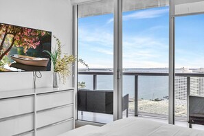 Un dormitorio moderno con un amplio ventanal que da a una masa de agua y edificios, una mesita de noche y una silla.