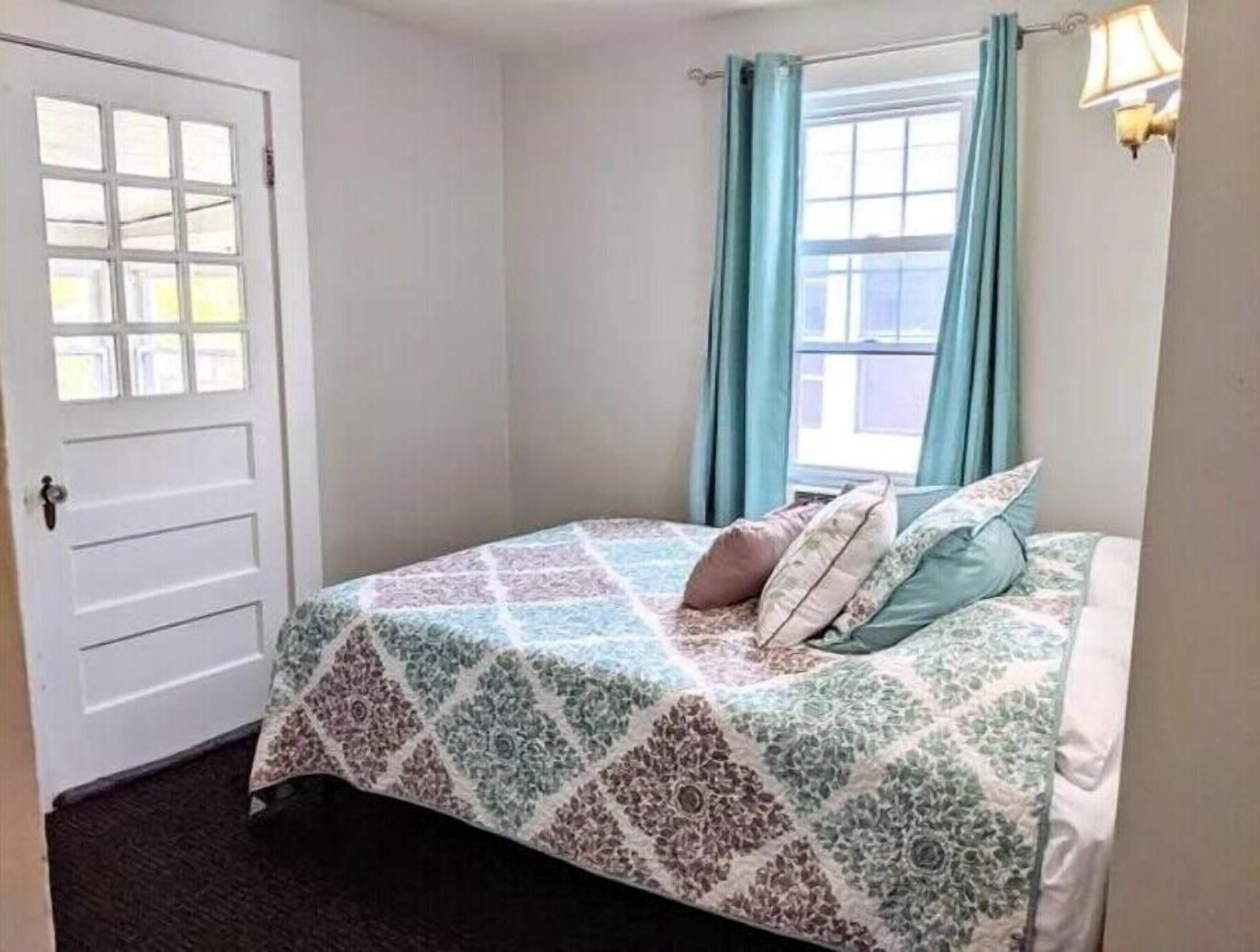 Un dormitorio con cama, ventana con cortinas, puerta y una lámpara fijada a la pared.